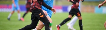 Two soccer players contest the ball on the field, one in a blue shirt and the other in a black and orange kit.